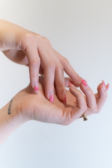 Two hands are positioned in a graceful, intertwined pose, displaying an artistic nail design with pink, flame-like patterns. A small tattoo is visible on the wrist, and a ring adorns one finger.