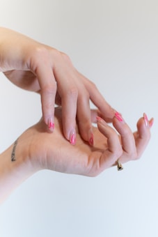 Two hands are positioned in a graceful, intertwined pose, displaying an artistic nail design with pink, flame-like patterns. A small tattoo is visible on the wrist, and a ring adorns one finger.