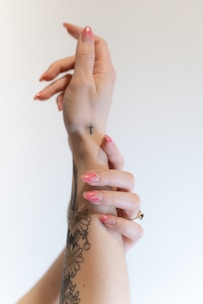 Hands are gracefully posed against a plain background. One arm displays a tattoo of intricate floral designs and a small cross tattoo on the wrist. The fingernails are painted with a glossy pink polish.