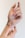 A close-up image of hands featuring a unique nail design with pink flame patterns over a nude base. The tattoo is visible on the wrist, and the hands are gracefully posed against a plain background.
