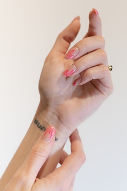 A close-up image of hands featuring a unique nail design with pink flame patterns over a nude base. The tattoo is visible on the wrist, and the hands are gracefully posed against a plain background.