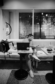 A person is sitting on a couch at a small table, looking at a smartphone with a laptop open in front of them. There are drinks and a bag nearby, inside a modern cafe with large windows showing reflections and possibly plants outside.