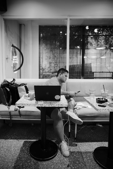 A person is sitting on a couch at a small table, looking at a smartphone with a laptop open in front of them. There are drinks and a bag nearby, inside a modern cafe with large windows showing reflections and possibly plants outside.