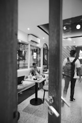 A black-and-white image of a modern café interior with a person sitting at a table with a laptop and drinks. Another person is walking away, carrying a backpack. The room has a comfortable seating arrangement with soft lighting and visible air conditioning units.