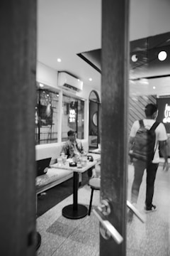 A black-and-white image of a modern café interior with a person sitting at a table with a laptop and drinks. Another person is walking away, carrying a backpack. The room has a comfortable seating arrangement with soft lighting and visible air conditioning units.