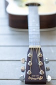 A close-up perspective of a guitar's headstock, showcasing tuning pegs, strings, and the brand name 'Martin Co.' engraved on a dark wooden surface. The blurred body of the acoustic guitar can be seen in the background, lying on a light wooden surface.