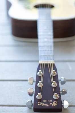 A close-up perspective of a guitar's headstock, showcasing tuning pegs, strings, and the brand name 'Martin Co.' engraved on a dark wooden surface. The blurred body of the acoustic guitar can be seen in the background, lying on a light wooden surface.