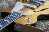 A close-up view of a guitar with a focus on the strings, switch, and part of the body. The guitar has a glossy wood finish and is placed on a textured surface, possibly straw. Details include the control knob and visible parts of the pickguard.