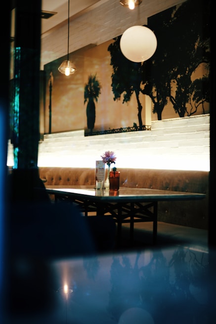 A cozy seating area in a dimly lit cafe or restaurant, featuring a table set against a cushioned bench. The tabletop has a flower vase and some menu cards. The wall behind is adorned with artistic silhouettes of trees and a street lamp. Soft, warm lighting creates an inviting ambiance.