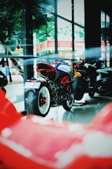 A motorcycle showroom featuring a stylish MV Agusta bike with a vibrant red framework and sleek design. The bike is positioned indoors with large windows that reflect outdoor scenery, including trees and buildings, creating a modern and energetic ambiance.