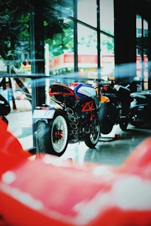 A motorcycle showroom featuring a stylish MV Agusta bike with a vibrant red framework and sleek design. The bike is positioned indoors with large windows that reflect outdoor scenery, including trees and buildings, creating a modern and energetic ambiance.