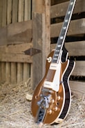A vintage-style electric guitar with a brown body and shiny metal components is resting against a wooden barn wall. The setting includes hay scattered on the ground, suggesting a rustic or country atmosphere.