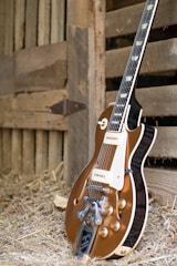 A vintage-style electric guitar with a brown body and shiny metal components is resting against a wooden barn wall. The setting includes hay scattered on the ground, suggesting a rustic or country atmosphere.