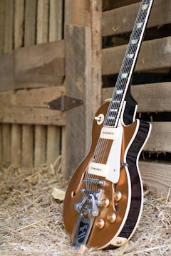 A vintage-style electric guitar with a brown body and shiny metal components is resting against a wooden barn wall. The setting includes hay scattered on the ground, suggesting a rustic or country atmosphere.