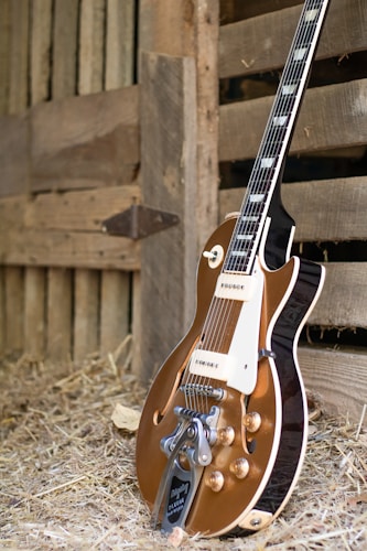 A vintage-style electric guitar with a brown body and shiny metal components is resting against a wooden barn wall. The setting includes hay scattered on the ground, suggesting a rustic or country atmosphere.