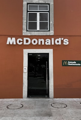A McDonald's restaurant entrance with a brown wall and a white-framed window above the door. The McDonald's logo is prominently displayed in white lettering, and there is a sign indicating accessibility. The entrance door is glass with a metallic frame.