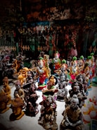 A collection of various intricately designed statues, including depictions of deities and spiritual figures, placed on display. The sculptures are crafted in a variety of colors and styles, with some figures playing musical instruments and others in prayerful poses. The background includes a metal fence and foliage, adding a natural, outdoor market setting.