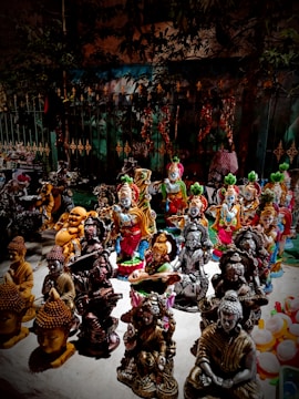 A collection of various intricately designed statues, including depictions of deities and spiritual figures, placed on display. The sculptures are crafted in a variety of colors and styles, with some figures playing musical instruments and others in prayerful poses. The background includes a metal fence and foliage, adding a natural, outdoor market setting.