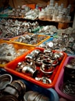 An assortment of stainless steel kitchenware is displayed in colorful plastic bins, arranged in a busy market setting. The items include small bowls, pots, and various kitchen utensils, tightly packed together. In the background, more kitchenware and items in plastic packaging are stacked on shelves.