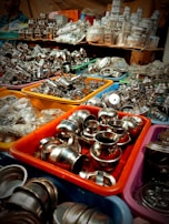 An assortment of stainless steel kitchenware is displayed in colorful plastic bins, arranged in a busy market setting. The items include small bowls, pots, and various kitchen utensils, tightly packed together. In the background, more kitchenware and items in plastic packaging are stacked on shelves.