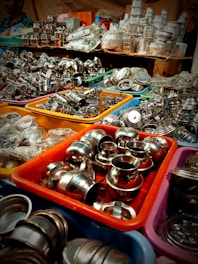 An assortment of stainless steel kitchenware is displayed in colorful plastic bins, arranged in a busy market setting. The items include small bowls, pots, and various kitchen utensils, tightly packed together. In the background, more kitchenware and items in plastic packaging are stacked on shelves.