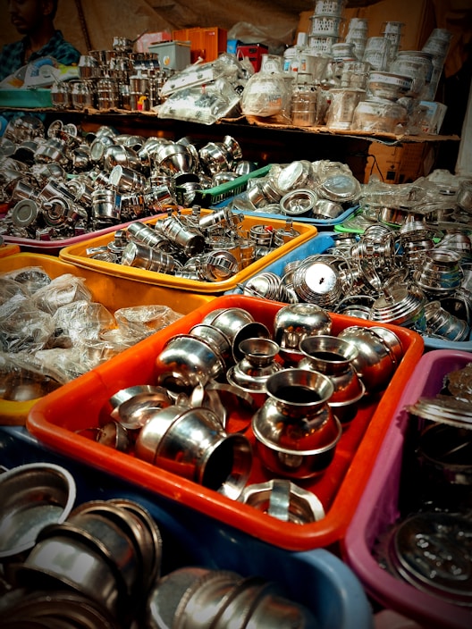 An assortment of stainless steel kitchenware is displayed in colorful plastic bins, arranged in a busy market setting. The items include small bowls, pots, and various kitchen utensils, tightly packed together. In the background, more kitchenware and items in plastic packaging are stacked on shelves.