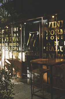 A cozy caf&eacute; setting with wooden tables and stools positioned outside a storefront featuring large glass windows. The glass is adorned with gold lettering, creating a warm, inviting atmosphere enhanced by soft lighting.