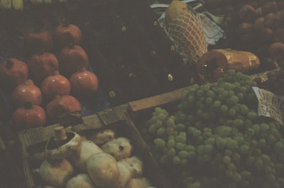 A market stall displaying a variety of fruits including pomegranates, grapes, and possibly breadfruit. The scene is dimly lit, giving an earthy and rustic atmosphere. A handwritten sign lists the price of some items. An item wrapped in yellow netting is visible in the background.