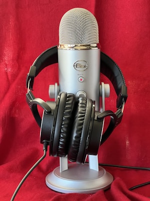 A large, silver microphone is prominently displayed with a pair of black headphones resting on its base. The background is a rich red fabric, creating a striking contrast with the metallic and black elements of the microphone and headphones.