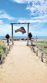 A sandy pathway lined with wooden posts and ropes leads to a beach, with clear blue skies and fluffy clouds overhead. Two hammocks are hanging from posts, with palm-like plants on either side. In the distance, a sign reads 'Caminho da Roça' in a rustic style. The ocean is visible in the background, with a small umbrella seen on the beach.