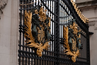An ornate wrought iron gate adorned with intricate golden embellishments featuring lions, crowns, and emblems. The craftsmanship is detailed, with black metal bars and decorative gold accents creating a regal and majestic appearance.