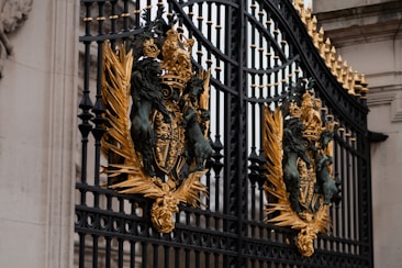 An ornate wrought iron gate adorned with intricate golden embellishments featuring lions, crowns, and emblems. The craftsmanship is detailed, with black metal bars and decorative gold accents creating a regal and majestic appearance.
