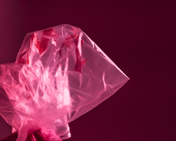 A translucent pink plastic bag with a crinkled texture, contrasting against a deep, maroon background. The lighting highlights the shiny surface and folds of the plastic, creating a dynamic play of light and shadow.