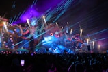A vibrant, colorful outdoor event at night features a large, elaborate stage with giant mythical female figures and intricate designs. Bright lasers and fireworks light up the sky, while smoke and whimsical, fantastical elements enhance the scene. A lively crowd of people is visible in the foreground, capturing the spectacle on their phones.
