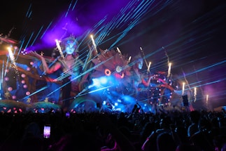 A vibrant, colorful outdoor event at night features a large, elaborate stage with giant mythical female figures and intricate designs. Bright lasers and fireworks light up the sky, while smoke and whimsical, fantastical elements enhance the scene. A lively crowd of people is visible in the foreground, capturing the spectacle on their phones.