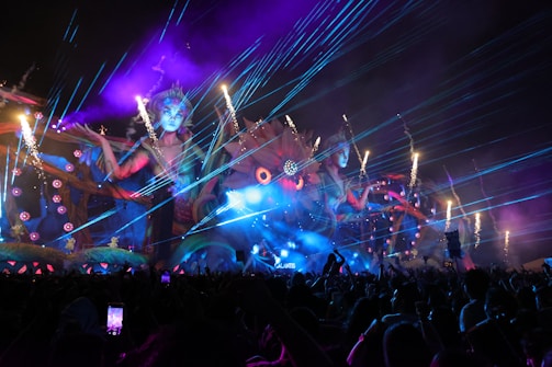 A vibrant, colorful outdoor event at night features a large, elaborate stage with giant mythical female figures and intricate designs. Bright lasers and fireworks light up the sky, while smoke and whimsical, fantastical elements enhance the scene. A lively crowd of people is visible in the foreground, capturing the spectacle on their phones.
