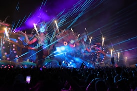 A vibrant, colorful outdoor event at night features a large, elaborate stage with giant mythical female figures and intricate designs. Bright lasers and fireworks light up the sky, while smoke and whimsical, fantastical elements enhance the scene. A lively crowd of people is visible in the foreground, capturing the spectacle on their phones.