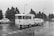A black and white image depicting a vintage car towing a caravan along a wet road. The scene is set outdoors, with trees lining the road and vintage street lamps visible. There are other classic cars on the road, and the ground appears wet, suggesting recent rain.