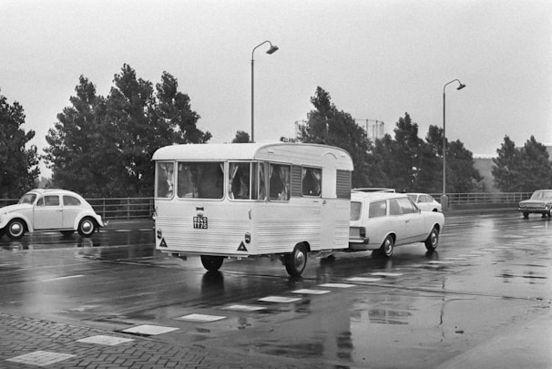 A black and white image depicting a vintage car towing a caravan along a wet road. The scene is set outdoors, with trees lining the road and vintage street lamps visible. There are other classic cars on the road, and the ground appears wet, suggesting recent rain.