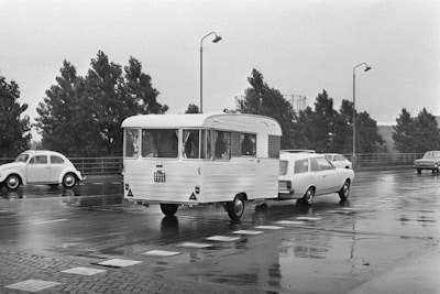 A black and white image depicting a vintage car towing a caravan along a wet road. The scene is set outdoors, with trees lining the road and vintage street lamps visible. There are other classic cars on the road, and the ground appears wet, suggesting recent rain.