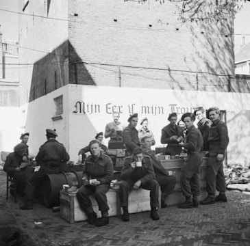 A group of soldiers are gathered in what appears to be an outdoor setting, possibly a courtyard with brick pavement and a wall with an inscription in Dutch. Some soldiers are seated while others are standing, holding cups and eating. The soldiers are wearing uniforms and berets.