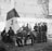 A group of soldiers are gathered in what appears to be an outdoor setting, possibly a courtyard with brick pavement and a wall with an inscription in Dutch. Some soldiers are seated while others are standing, holding cups and eating. The soldiers are wearing uniforms and berets.