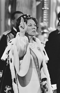 A woman dressed in ceremonial attire, including a robe with fur trim and a decorative headpiece, raises her hand in a gesture. She appears to be participating in a formal event, likely involving a significant cultural or historical context. Other figures are visible in the blurred background.