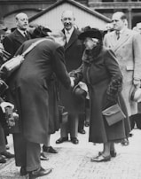 A group of formally dressed individuals in a vintage setting, with a woman in a coat and hat shaking hands with a man who is bowing slightly. Several others stand watching the interaction, including men in suits and overcoats. A person is visible holding a camera in the foreground.