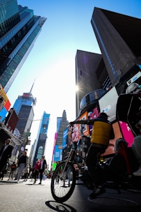 A bustling urban environment with tall skyscrapers surrounding a busy street. Various pedestrians are walking, while a person is riding a bicycle. Large digital billboards display advertisements, contributing to the vibrant atmosphere. The sun shines brightly, casting shadows and illuminating the scene.