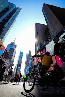 A bustling urban environment with tall skyscrapers surrounding a busy street. Various pedestrians are walking, while a person is riding a bicycle. Large digital billboards display advertisements, contributing to the vibrant atmosphere. The sun shines brightly, casting shadows and illuminating the scene.