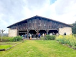 A group of people stands gathered in front of a large, rustic wooden barn with a weathered roof. The barn is surrounded by a vibrant green lawn with patches of yellow flowering plants. The sky is overcast with scattered clouds, creating a serene and peaceful rural scene.