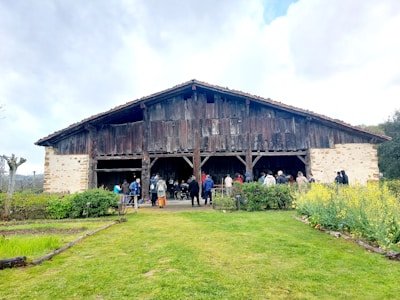 A group of people stands gathered in front of a large, rustic wooden barn with a weathered roof. The barn is surrounded by a vibrant green lawn with patches of yellow flowering plants. The sky is overcast with scattered clouds, creating a serene and peaceful rural scene.