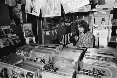 Two people are standing in a record store, flipping through vinyl records. Various music genres and album covers are visible, organized in rows. The space has a nostalgic, retro feel, with dim lighting and posters on the walls.