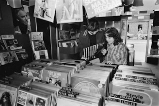 Two people are standing in a record store, flipping through vinyl records. Various music genres and album covers are visible, organized in rows. The space has a nostalgic, retro feel, with dim lighting and posters on the walls.